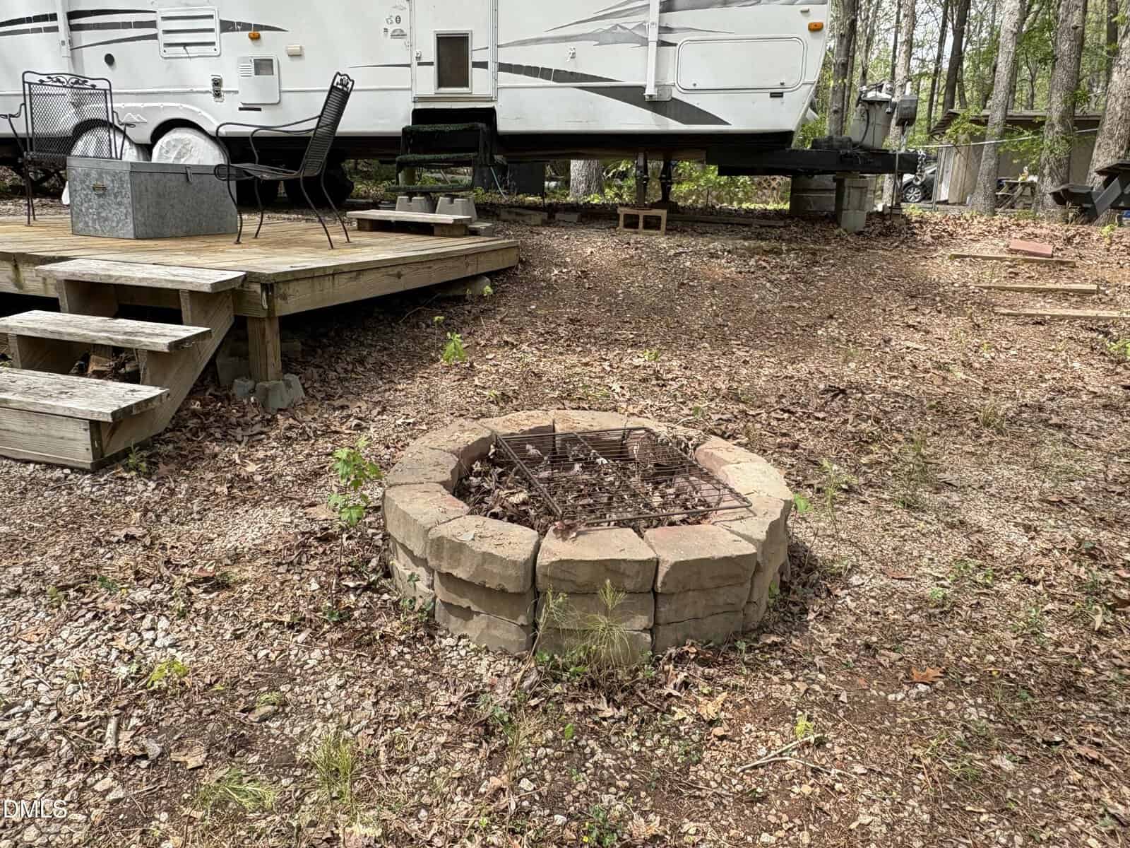 A round stone fire pit filled with leaves sits on dirt and gravel near a wooden deck with patio chairs. A large white RV is parked in the background among trees.