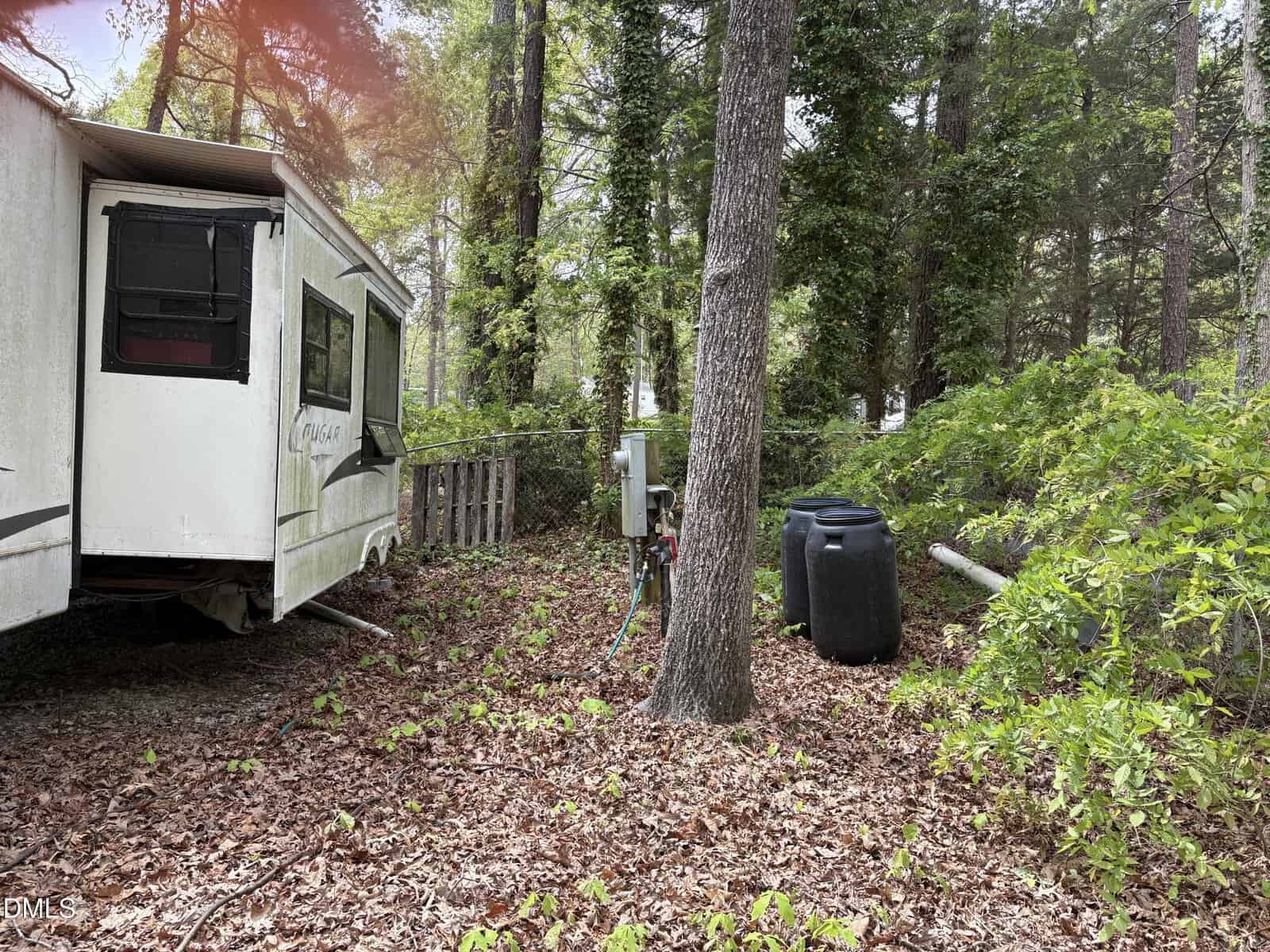 A white RV is parked in a wooded area with trees, green bushes, and fallen leaves on the ground. Two large black barrels and a gray utility box are situated near the RV, beside a tree.