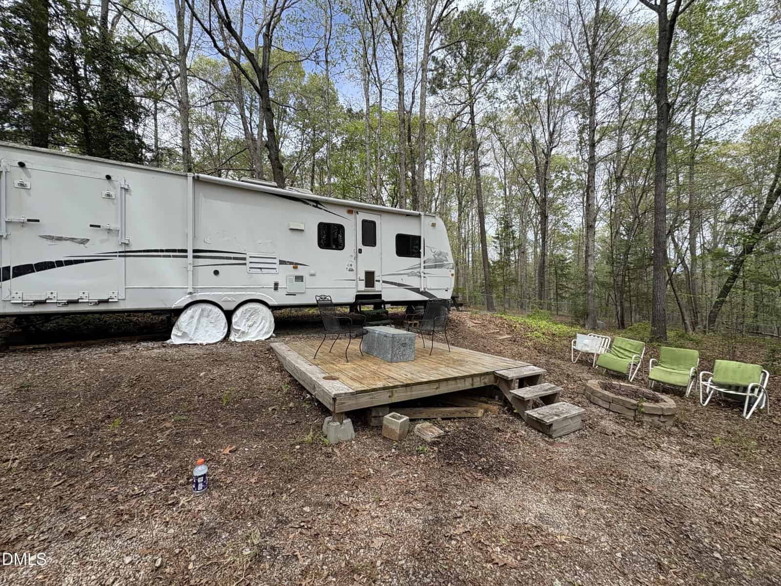 A white camper trailer is parked next to a wooden deck with three chairs and a table. Three green lounge chairs sit on the dirt beside the deck. The scene is in a wooded area with tall trees and scattered leaves.