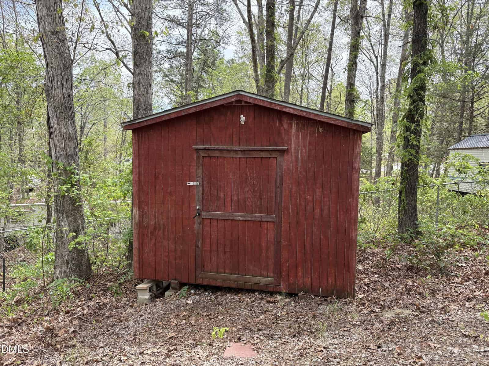 A small red wooden shed with a single door sits on uneven ground in a wooded area. Trees and leaves surround the shed, and a few concrete blocks are stacked near its base.