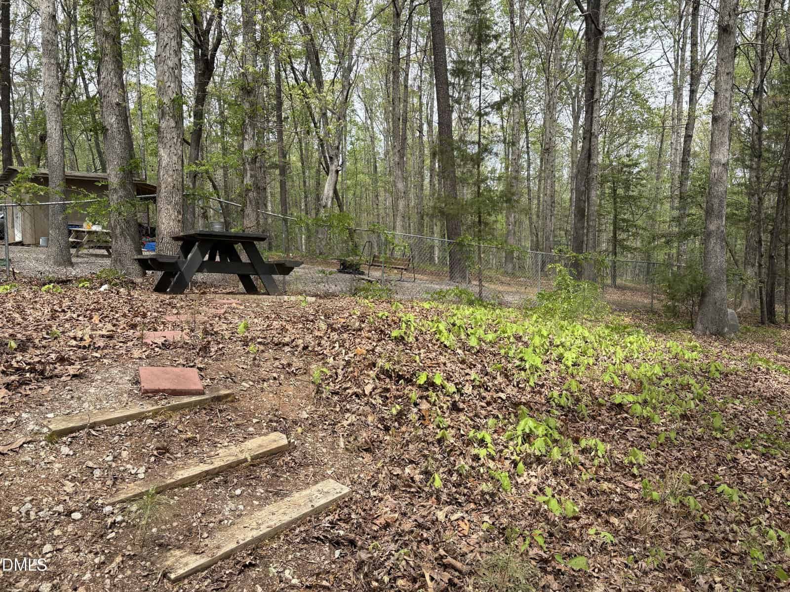 A wooded backyard with a black picnic table, wooden steps leading up a slope, a chain-link fence, and scattered leaves on the ground. A partial structure is visible on the left side.