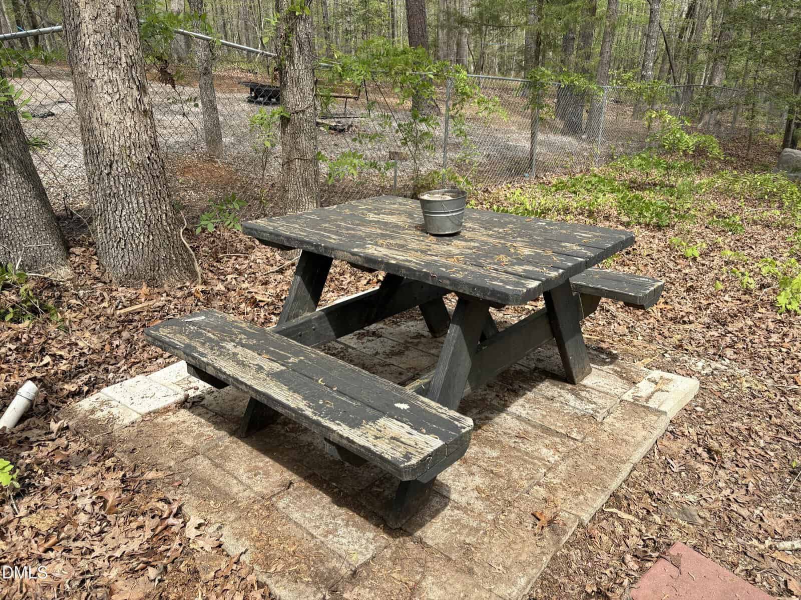 A weathered black wooden picnic table with attached benches sits on a patio surrounded by fallen leaves and trees. A small metal pot is placed on top of the table. A chain-link fence is visible in the background.