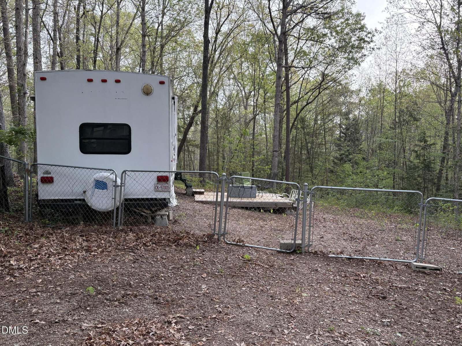 A white RV is parked next to a chain-link fence with a gate in a wooded area. A wooden deck with patio furniture sits nearby among tall trees and scattered leaves on the ground.