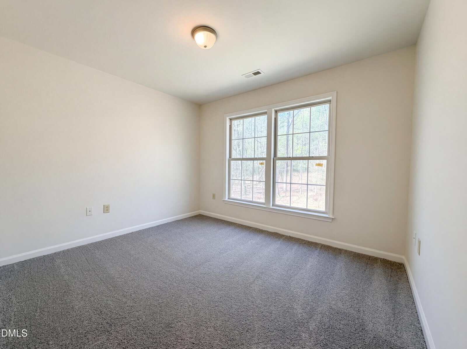 An empty room with light beige walls and grey carpet. A large window on the right wall provides a view of a wooded area outside. A single flush-mount light fixture is on the white ceiling. The room includes several electrical wall outlets.