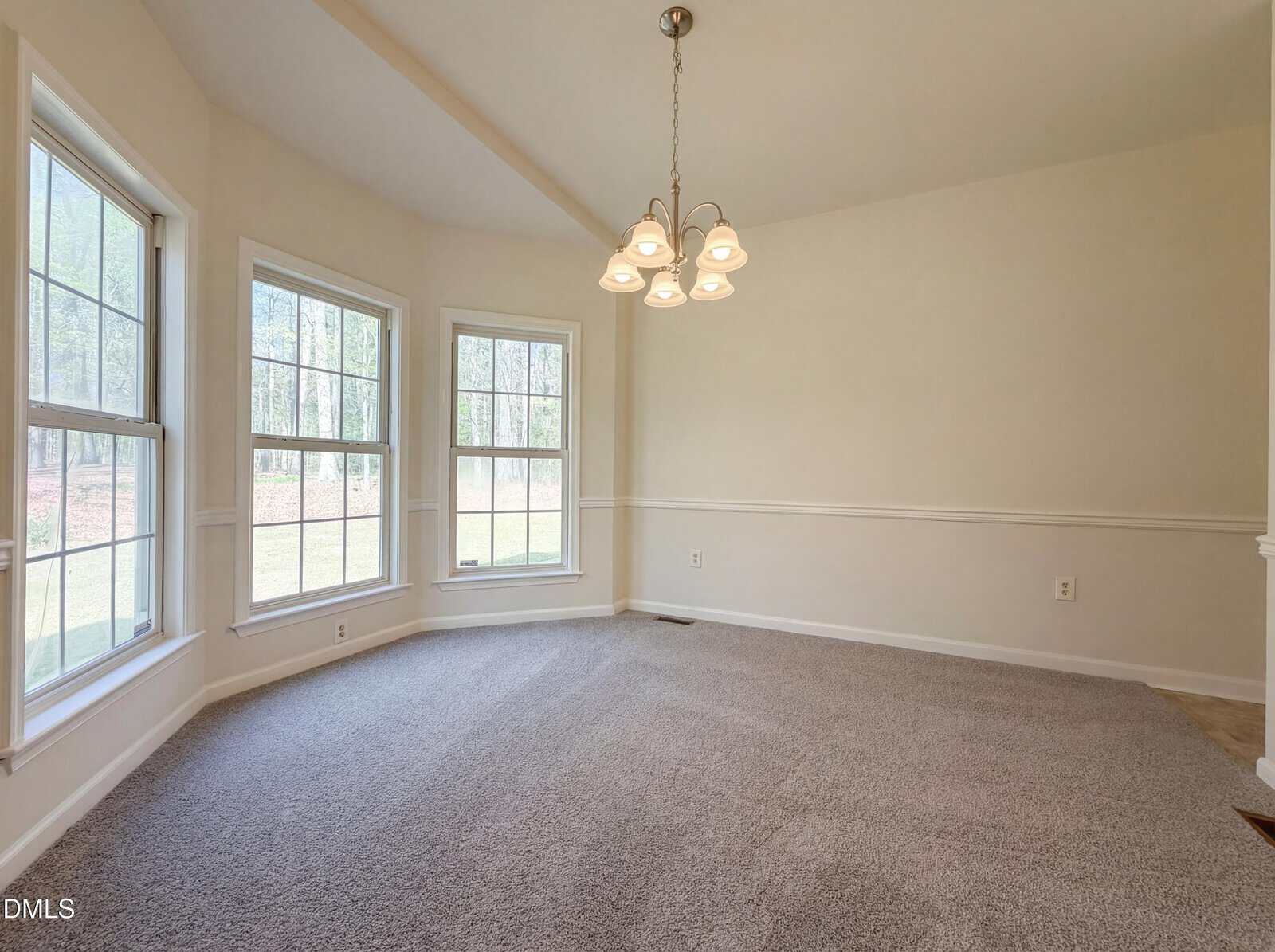 A carpeted dining area with beige walls, three large windows allowing natural light, a white chair rail detail, and a hanging light fixture with five bulbs. The room is empty and overlooks a grassy yard.