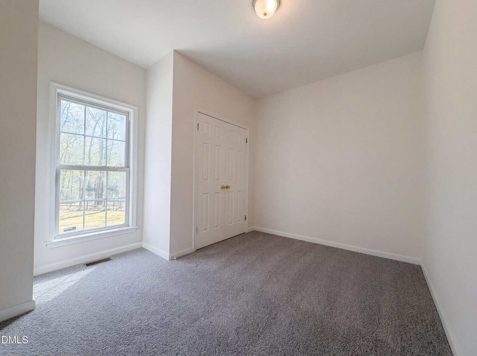 Empty room with light gray carpet, white walls, a double-door closet, ceiling light, and a large window letting in natural light.