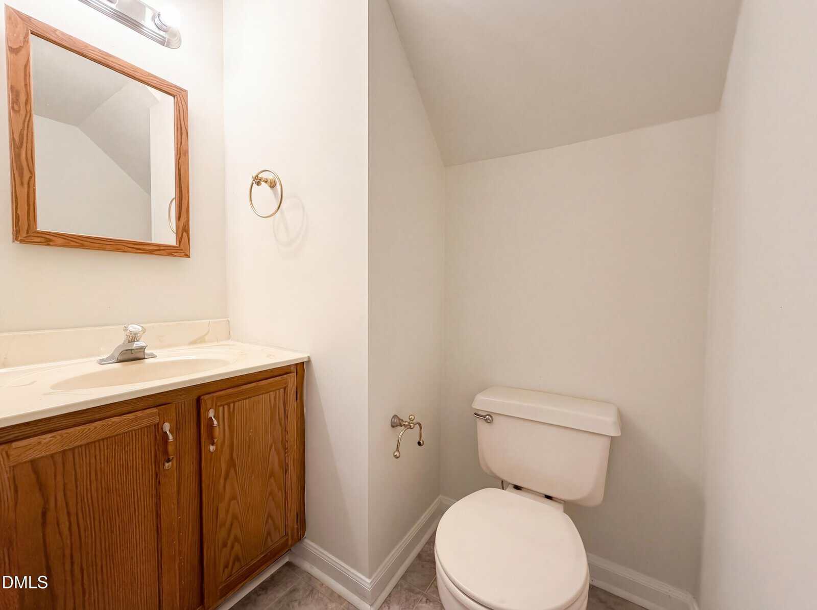 A small bathroom with a wooden vanity and sink on the left, a mirror above, and a white toilet on the right beneath a sloped ceiling. The walls and floor are light-colored.