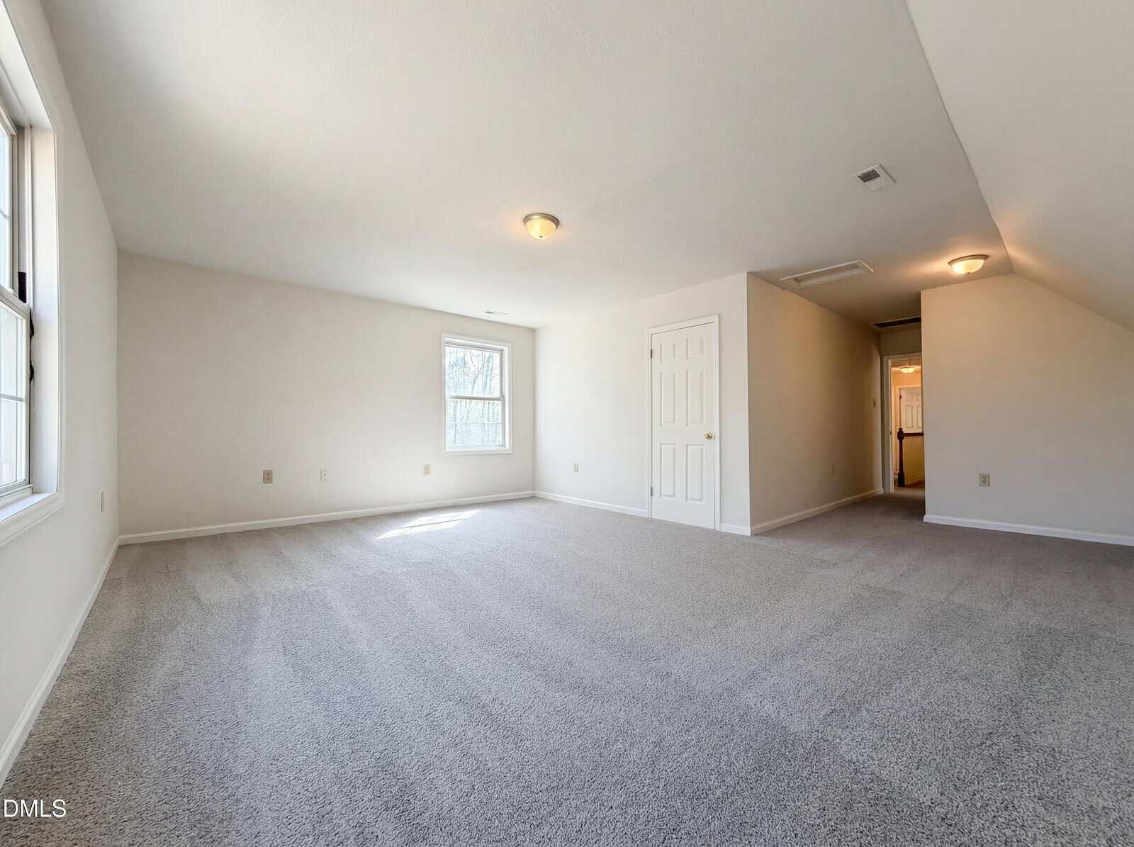 A spacious, empty room with light-colored walls and carpeted flooring. Two windows let in natural light, and there are ceiling lights. A white double door and a hallway leading to another room are visible.