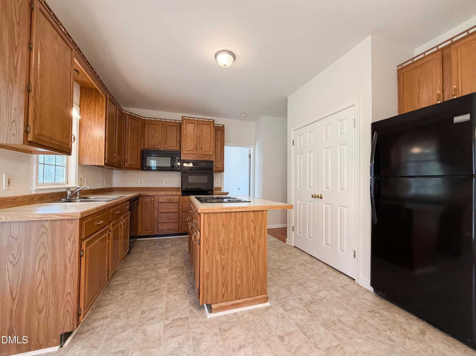 A kitchen with wood cabinets, a center island, black appliances including a refrigerator and microwave, double doors to a pantry, and a window above the sink. The floor has a light-colored tile pattern.