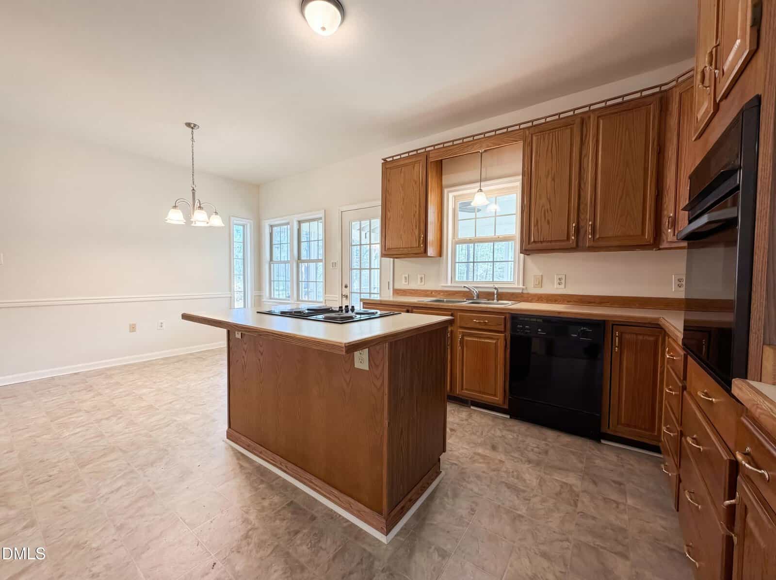 Spacious kitchen with wood cabinets, central island, black appliances, and large windows. Adjacent dining area with hanging light fixture and glass doors leading outside. Neutral tile flooring throughout.
