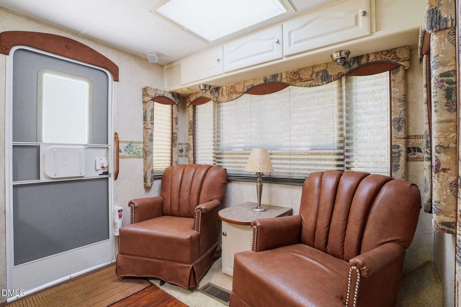 Two brown leather armchairs sit next to a round table with a lamp in front of a window with blinds in a cozy RV interior. The door is to the left, and patterned curtains frame the windows.
