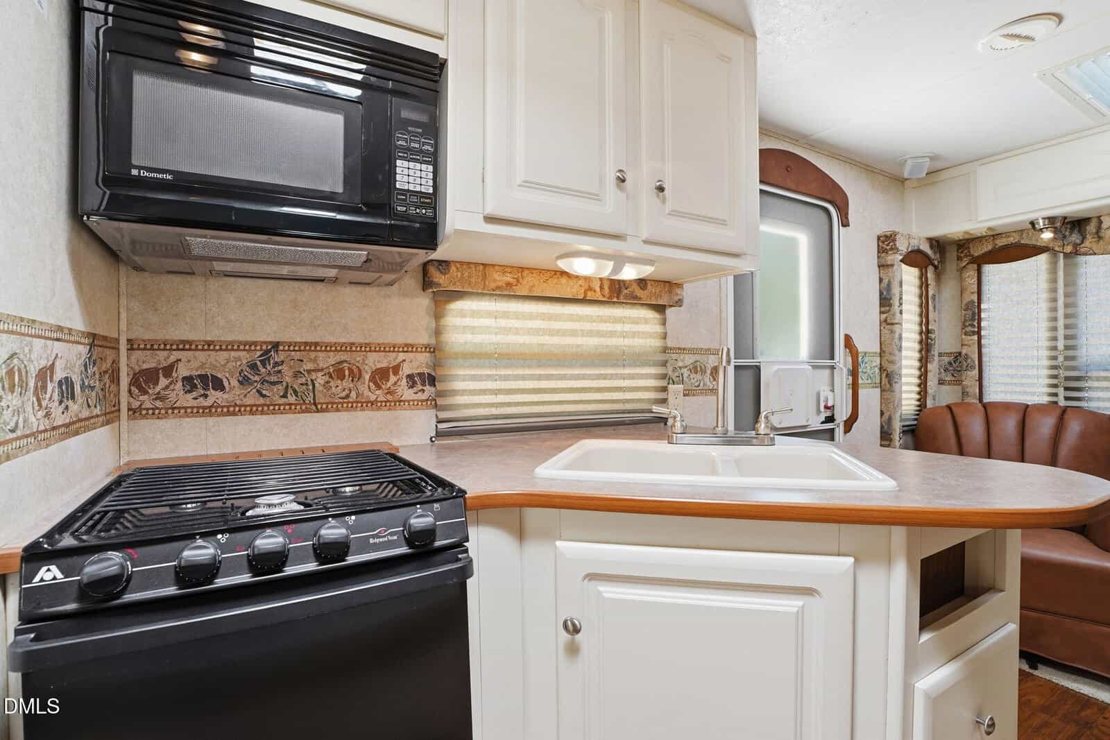 Compact RV kitchen with a black stove, oven, and microwave, white cabinets, double sink, and beige countertops. Light brown patterned backsplash and a window with a beige shade are visible above the sink.