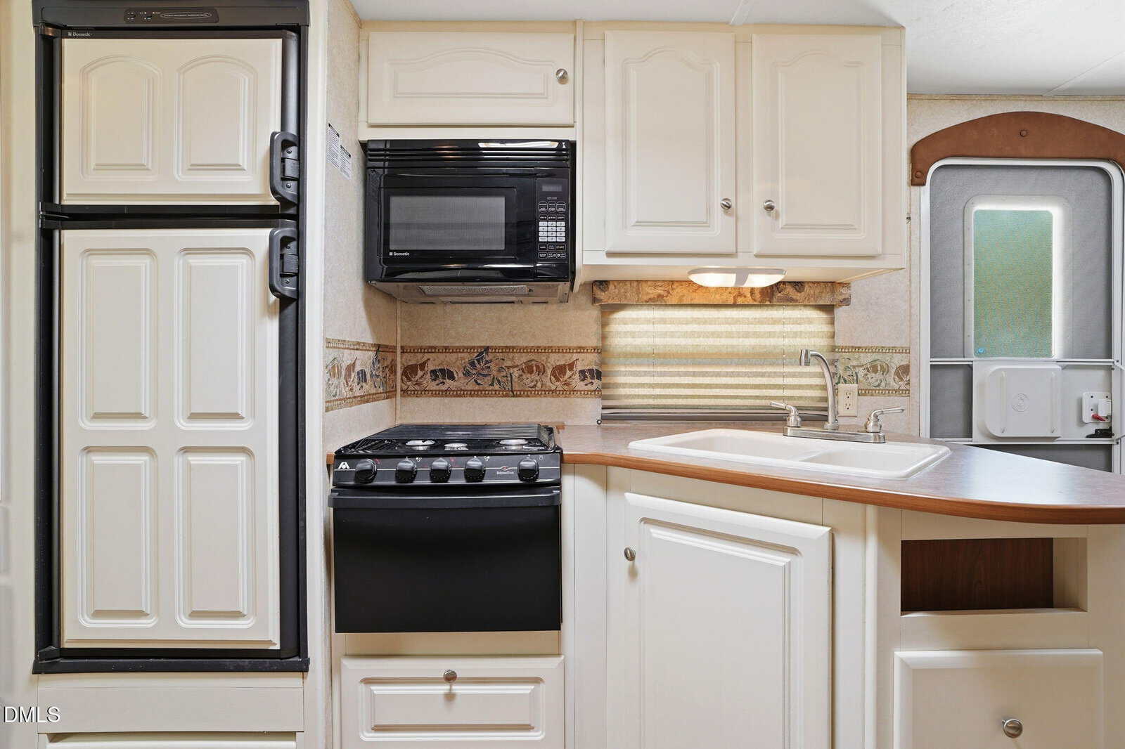 Compact RV kitchen with white cabinets, a black stove and microwave, a small fridge, and a single-basin sink with a faucet. Light under cabinets, striped backsplash, and a door visible on the right.