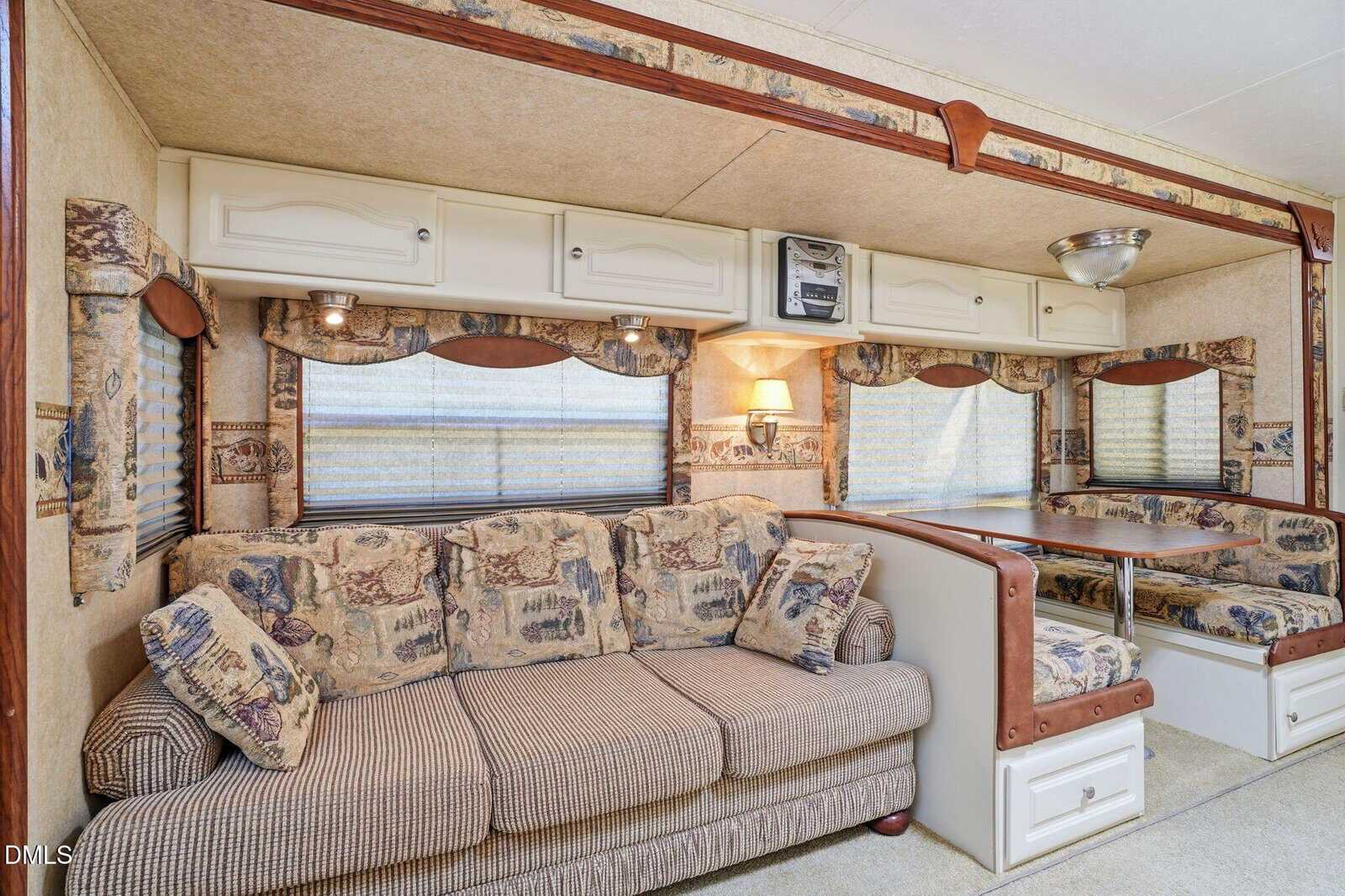 Interior of an RV with a beige patterned sofa, overhead cabinets, and a dining area featuring cushioned bench seating and a table, all decorated in a rustic, cabin-like style.