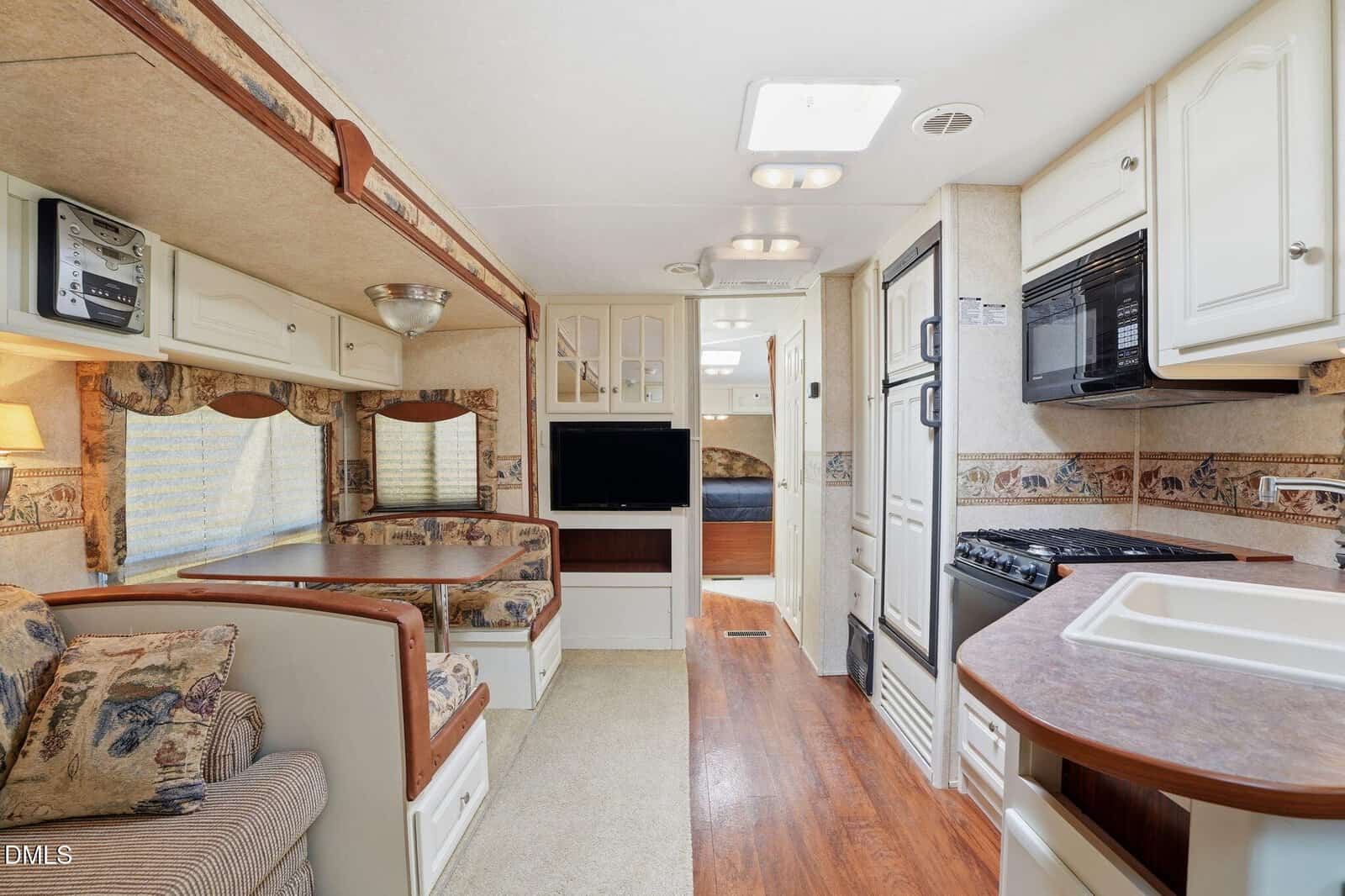 Interior of an RV with a kitchen area on the right, including a stove, microwave, and sink, and a dining booth and couch on the left. There is a TV in the center and a bed visible in the back.