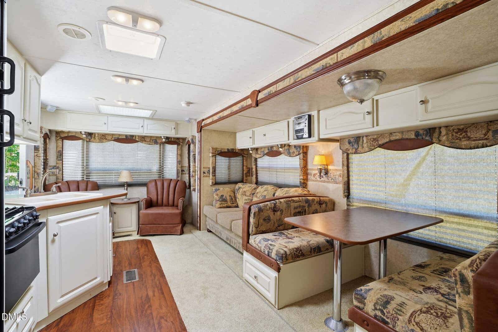 Interior of an RV with a kitchen, brown cushioned sofa, matching dining area, armchair, and large windows with patterned valances and blinds. Wood and beige tones create a cozy, compact living space.
