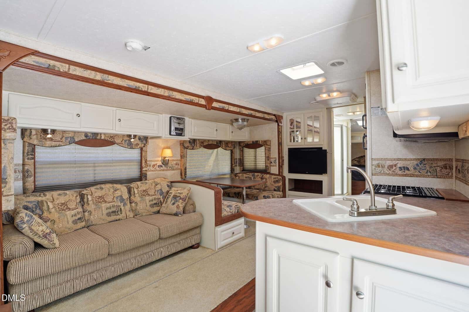 Interior of a well-lit RV featuring a beige sofa with patterned cushions, a small dinette, white cabinetry, a kitchen area with a sink, and a wall-mounted TV. Neutral decor and ample natural light.