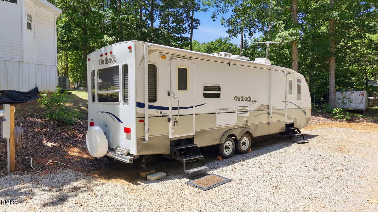 A white Outback travel trailer with two axles is parked on a gravel lot at 219 Eagle Drive in Louisburg, next to a white house and surrounded by trees and greenery on a sunny day.