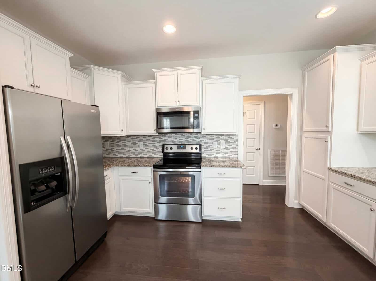 Modern kitchen with white cabinets, stainless steel appliances including a refrigerator, oven, and microwave, mosaic tile backsplash, granite countertops, and dark hardwood floors. Brightly lit with recessed ceiling lights.