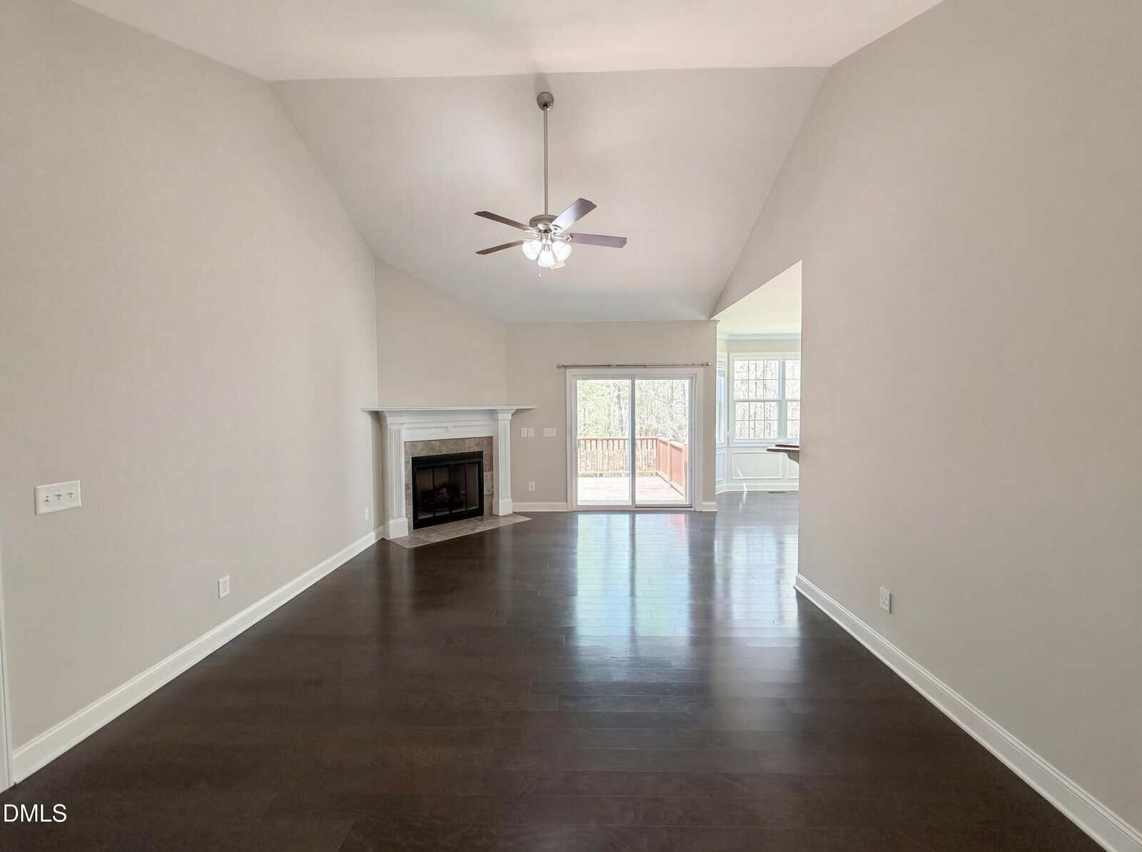 Spacious, empty living room with dark wood floors, light-colored walls, a white fireplace on the left, ceiling fan, and sliding glass doors leading to a deck with natural light streaming in.