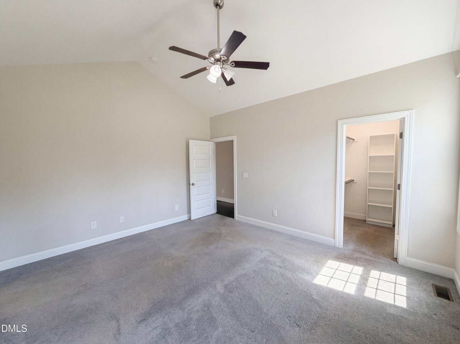 Bright, empty bedroom with gray carpet, pale walls, ceiling fan, door to walk-in closet, and sunlight streaming through a window creating a square of light on the floor.