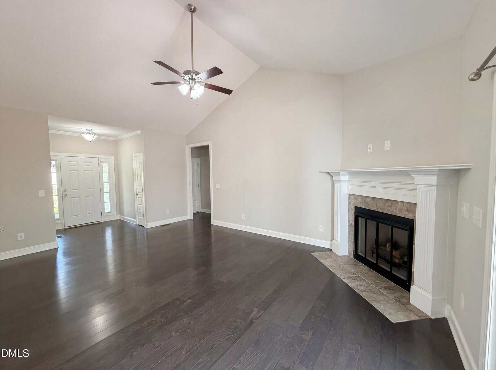 A spacious, empty living room with dark wood floors, vaulted ceiling with two ceiling fans, a white mantel fireplace, neutral-colored walls, and an entryway visible in the background.