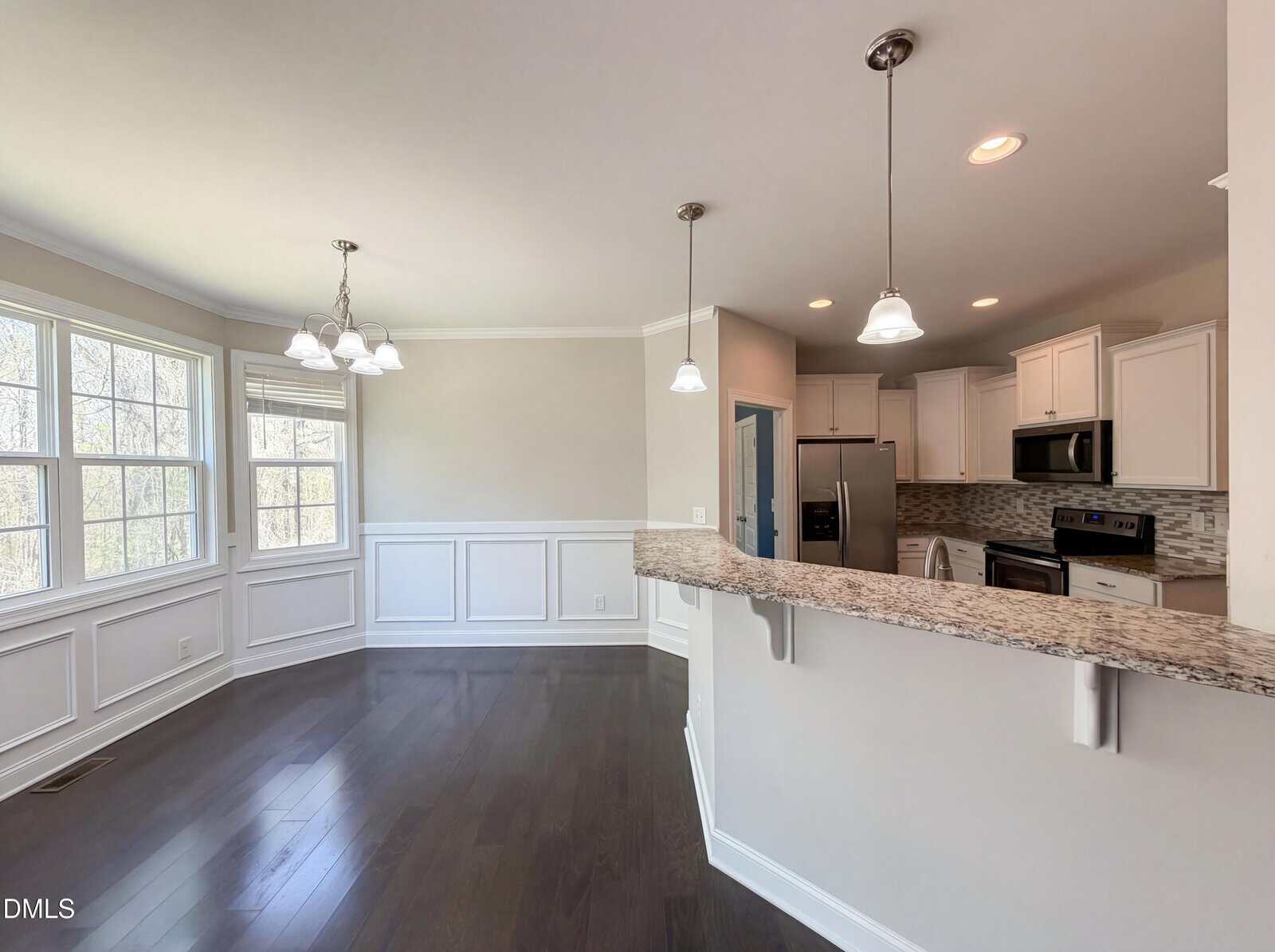 Bright kitchen with granite countertops, stainless steel appliances, dark hardwood floors, and a breakfast nook with large windows and wainscoting. Three pendant lights hang over the counter.
