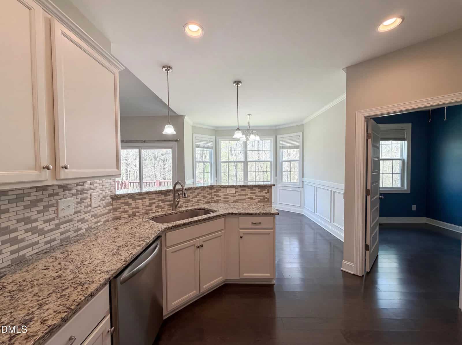 Modern kitchen with white cabinets, granite countertops, stainless steel appliances, and a tiled backsplash. A bay window and pendant lights brighten the adjacent dining area, with a view into a room with blue walls on the right.