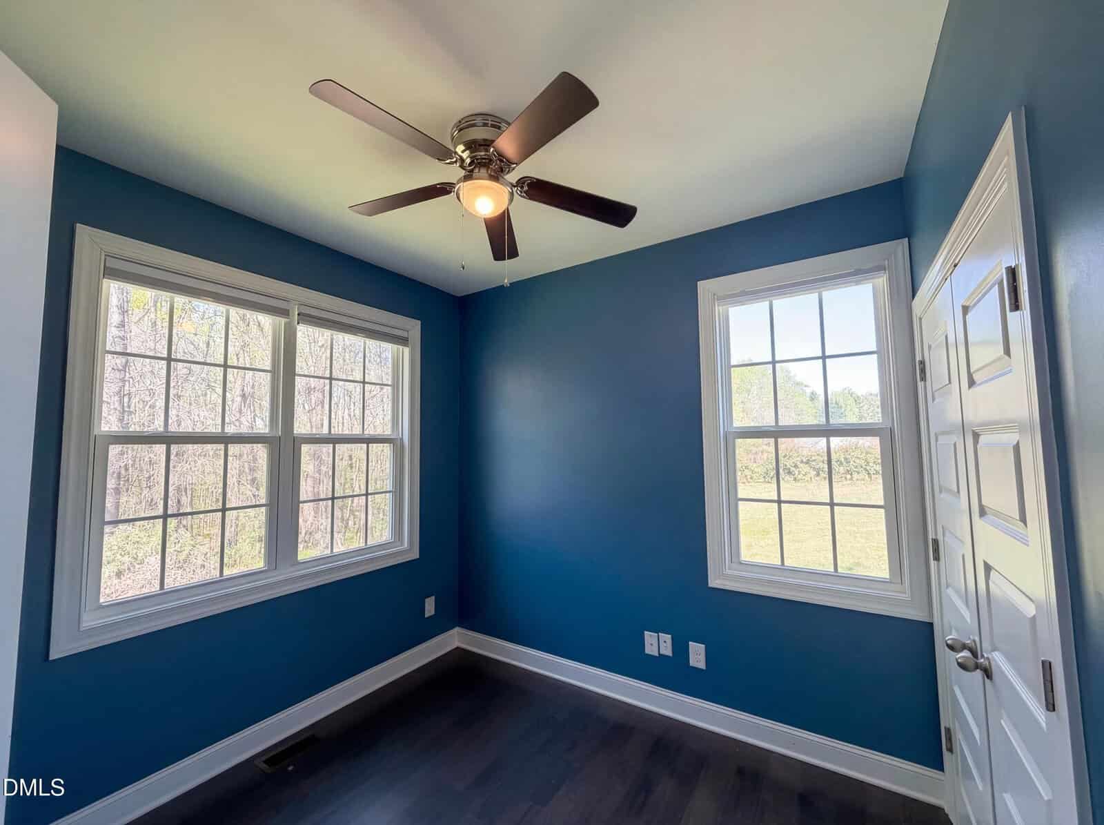 Small room with blue walls, two large windows letting in natural light, dark wood floor, white trim, and a ceiling fan with blades extended. There is a closed white door to the right side of the image.