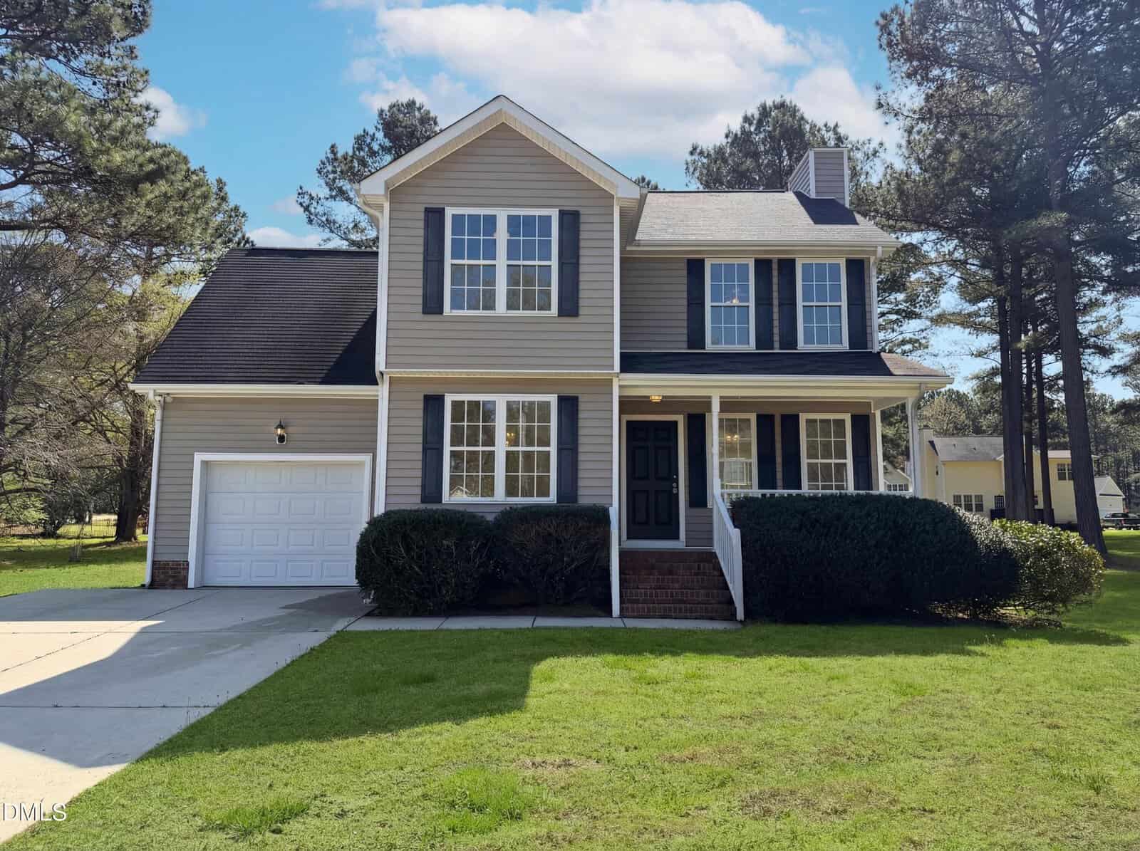 A two-story house with beige siding, white trim, and black shutters, featuring a covered front porch, attached single-car garage, and a well-maintained lawn bordered by bushes and trees.