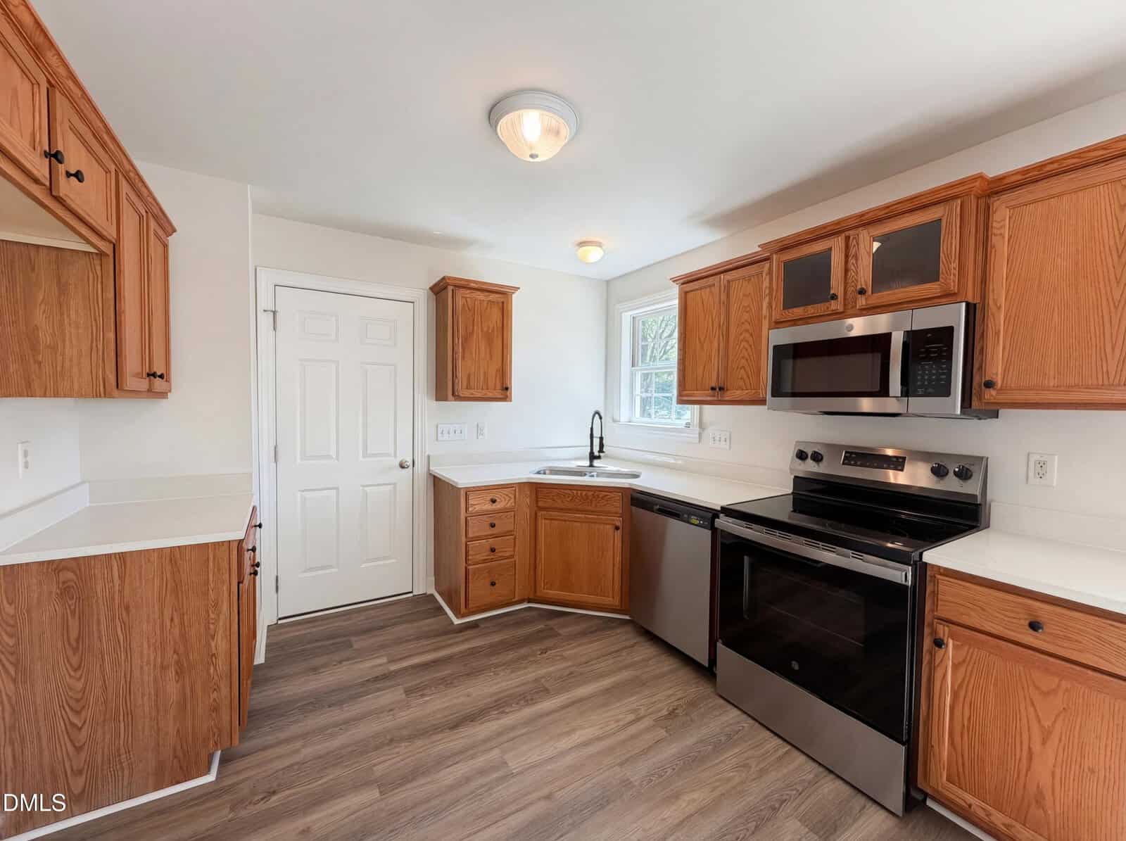Modern kitchen with wood cabinets, stainless steel appliances including a stove, microwave, and dishwasher, white countertops, and light wood flooring. Two windows let in natural light; overhead lighting is present.