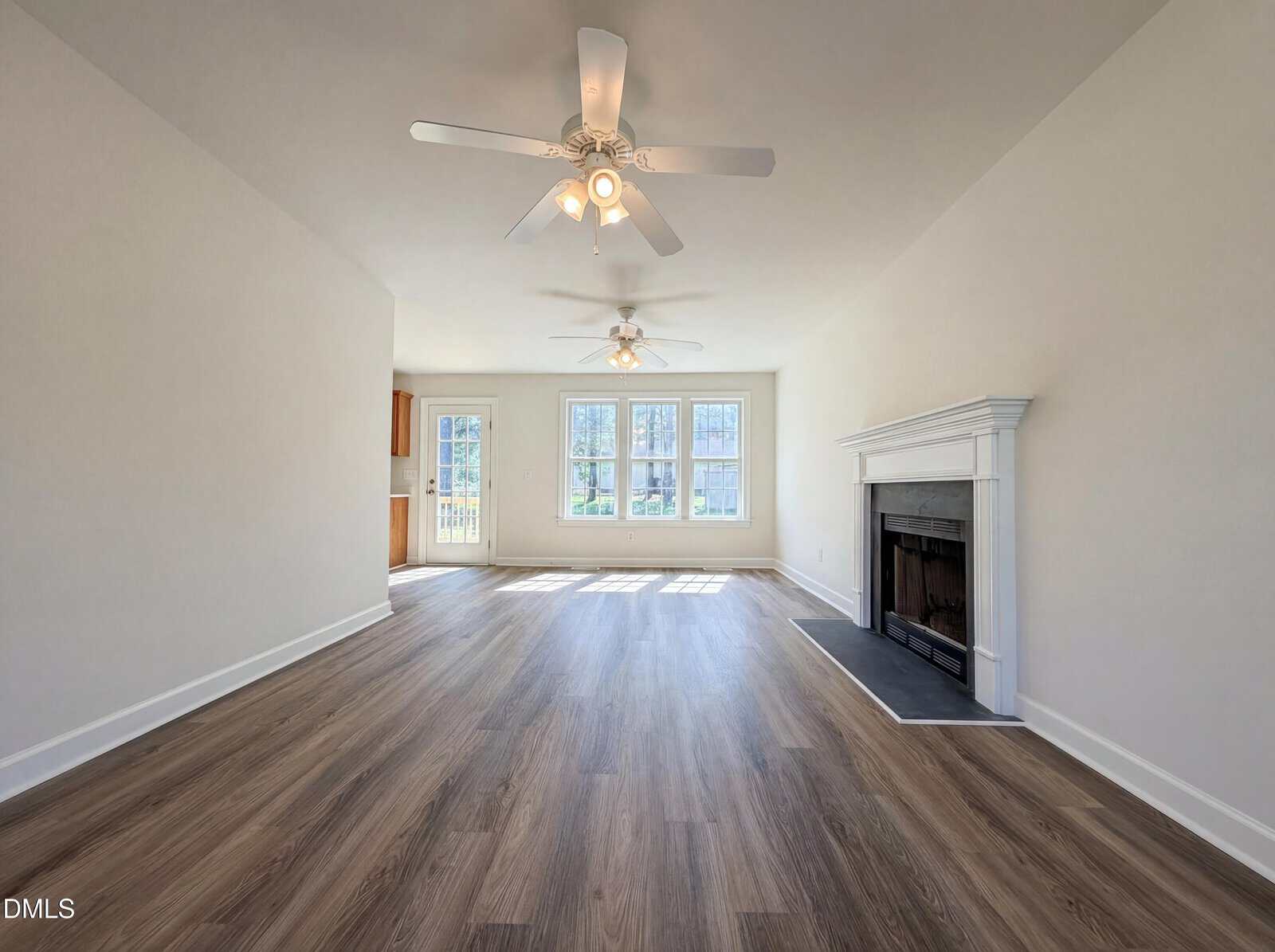 A spacious, empty living room with light-colored walls, wood flooring, two ceiling fans, large windows letting in natural light, and a white fireplace on the right wall.