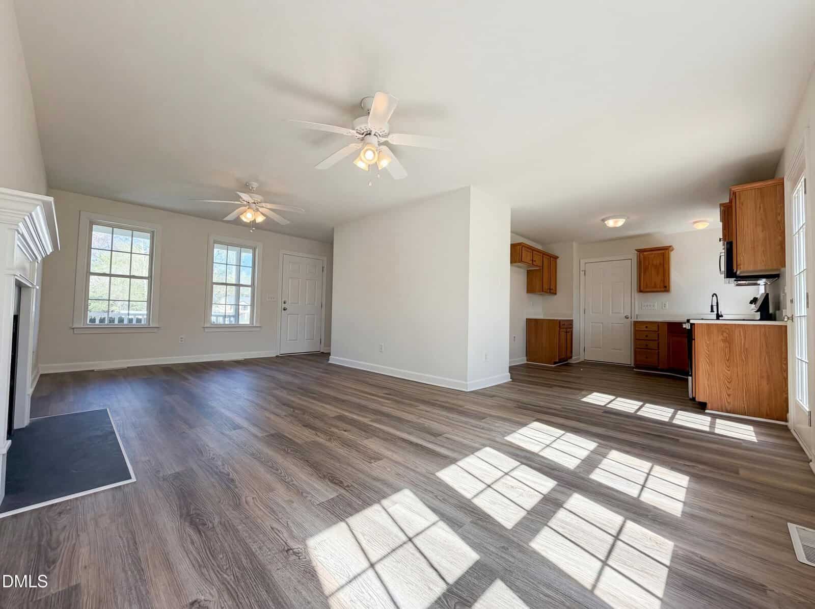 A bright, unfurnished open-concept living and kitchen area with wood-style flooring, white walls, ceiling fans, and large windows letting in natural light. Kitchen cabinets are wood-toned with white appliances.