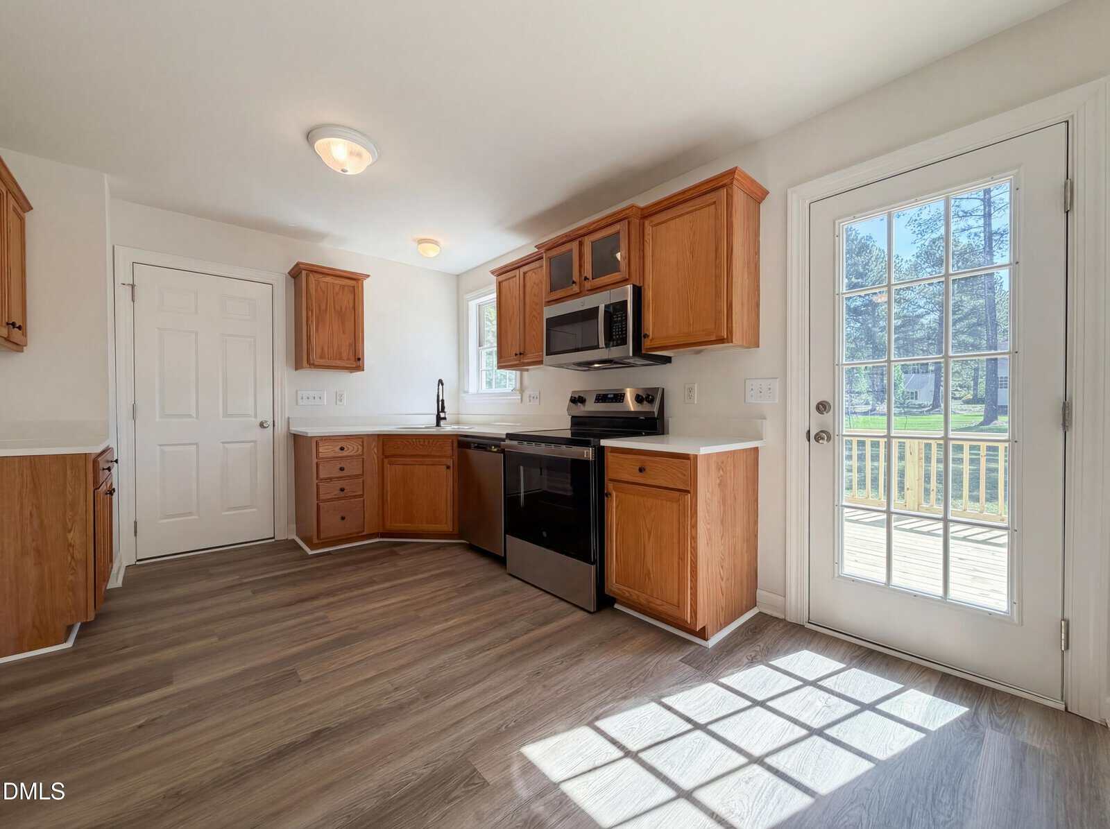 Bright kitchen with wood cabinets, black appliances, and light wood flooring. Sunlight streams through a glass door leading to a deck, creating a warm and inviting atmosphere. A window is above the sink.