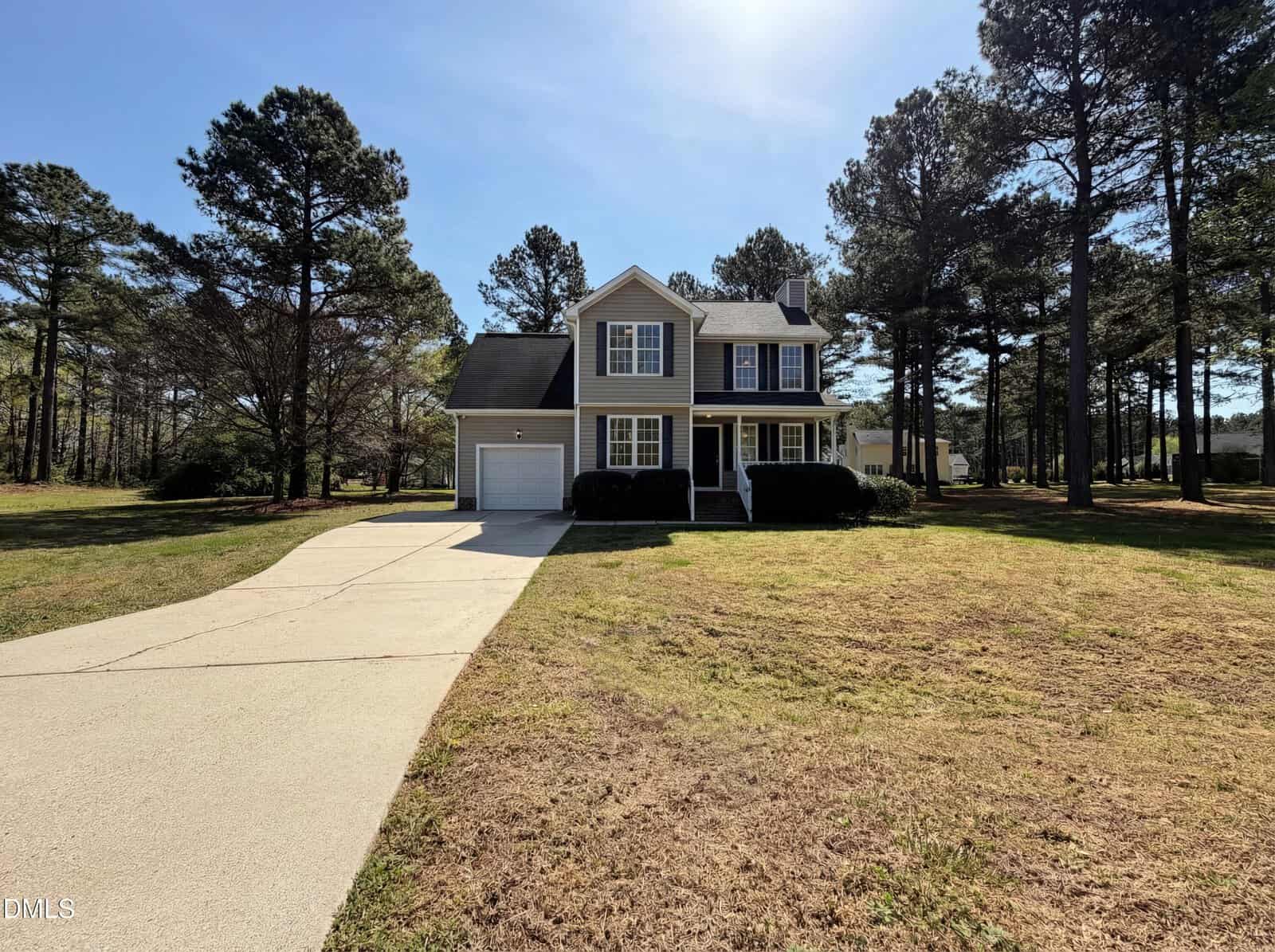 A two-story house with beige siding, white trim, and a front porch sits on a large lot with tall trees and a wide driveway leading to an attached single-car garage. The lawn is patchy with sunlight filtering through the trees.