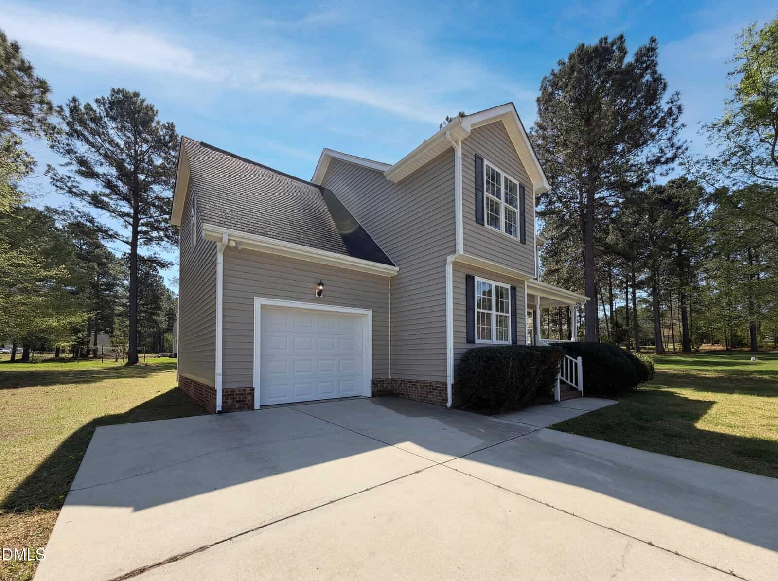 Two-story beige house with a single-car garage, white trim, and a covered front porch, set on a spacious lot with a concrete driveway, surrounded by grass, trees, and a clear sky.