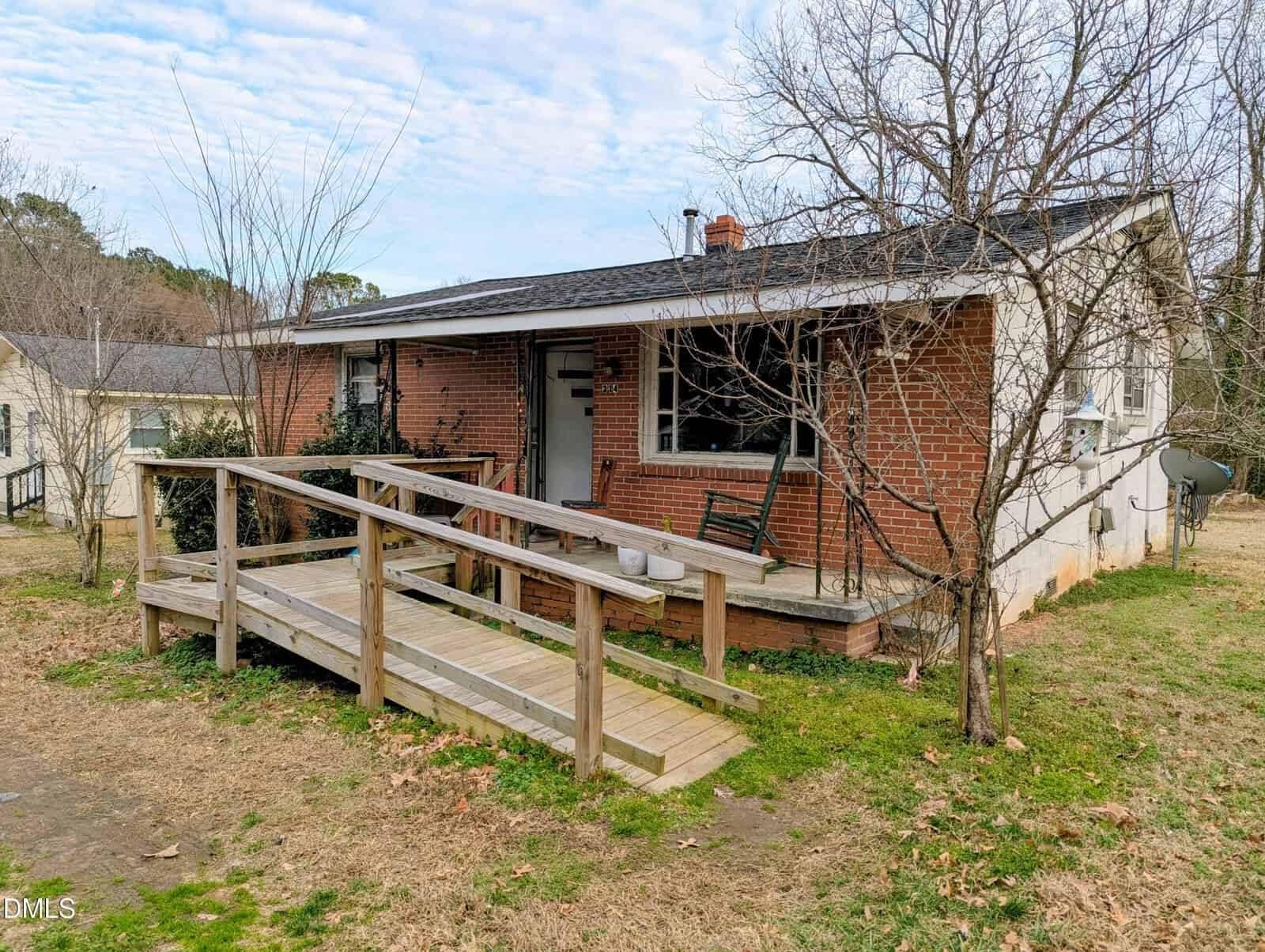 A small, single-story brick house with a covered porch, wheelchair-accessible wooden ramp, leafless trees, and patchy grass in the yard. A black rocking chair sits on the porch.