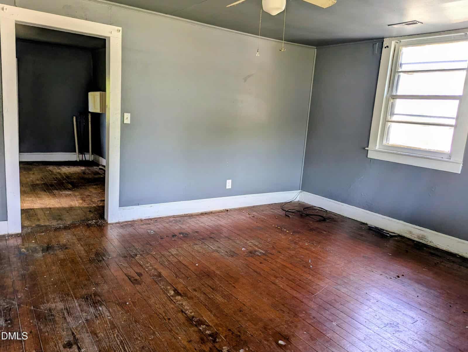 Empty room with faded gray walls, a ceiling fan, a window with white trim, and worn hardwood floors. An open doorway leads to another room with visible dirt and an old utility sink in the corner.