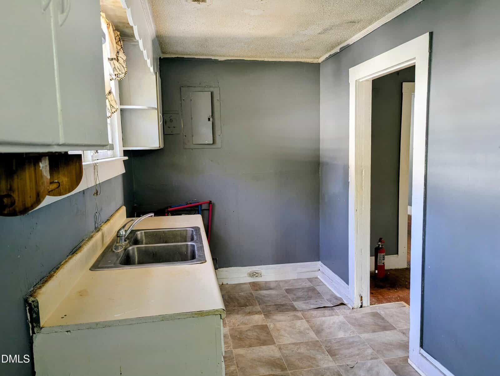 Small, empty kitchen with gray walls, white cabinets, a double sink, and a window. The floor is tiled, and a fire extinguisher is visible through the open doorway into the next room.