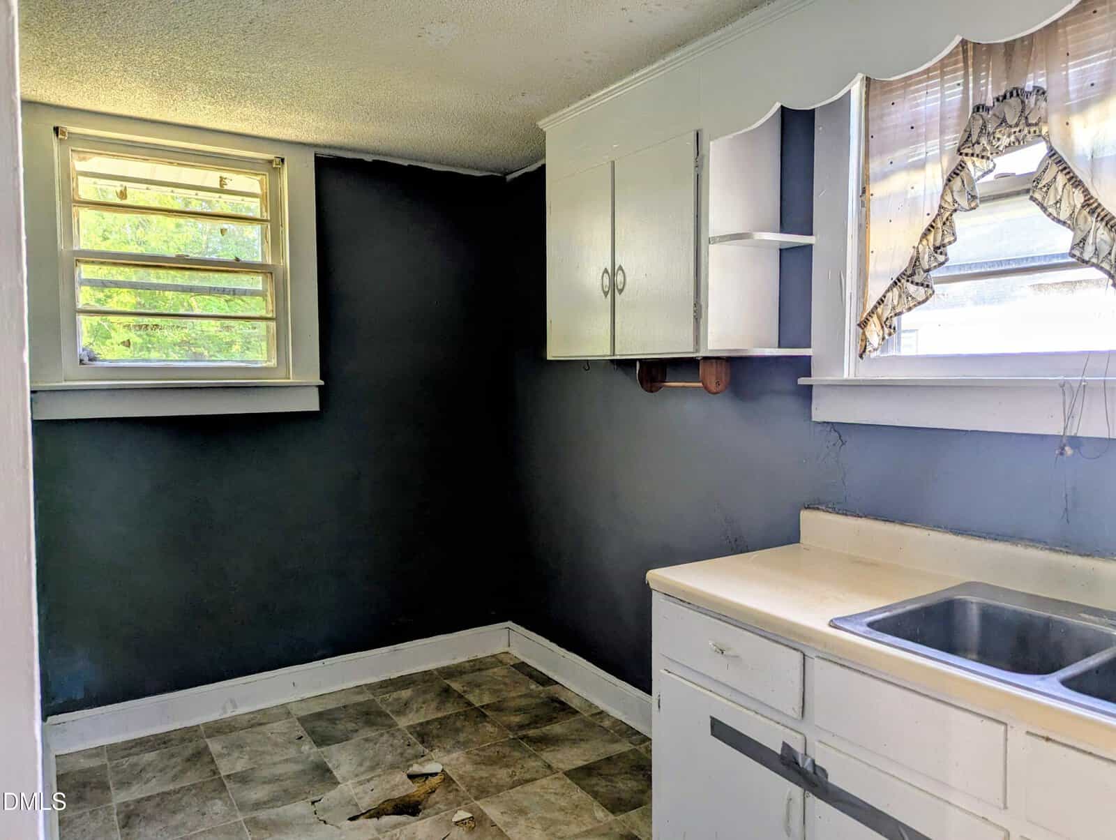 Small, empty kitchen with worn linoleum floor, white cabinets, a sink, and two windows with partial curtains. The walls are painted dark gray, and one section of the floor is damaged.