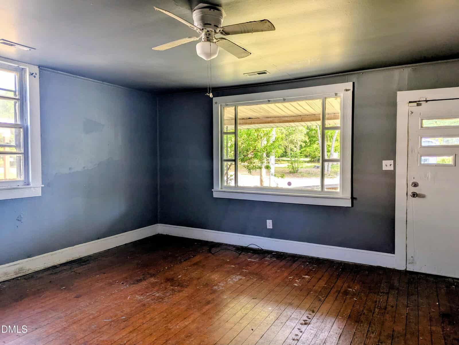 An empty room with dark blue walls, a white ceiling fan, wooden floor, two windows letting in natural light, and a white door with glass panels. The floor shows signs of wear and scuff marks.