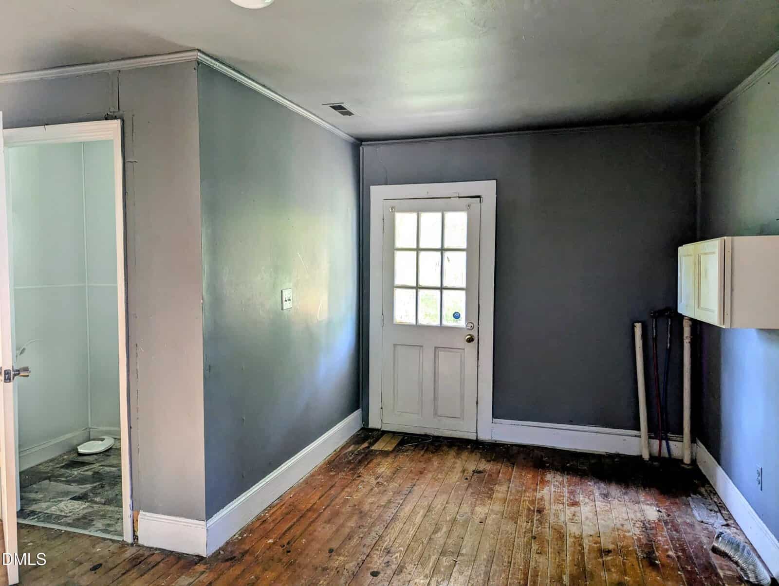 A small, empty room with gray walls, a worn wooden floor, a white door with glass panes, a wall vent, some exposed pipes, and an open doorway leading to a tiled bathroom.