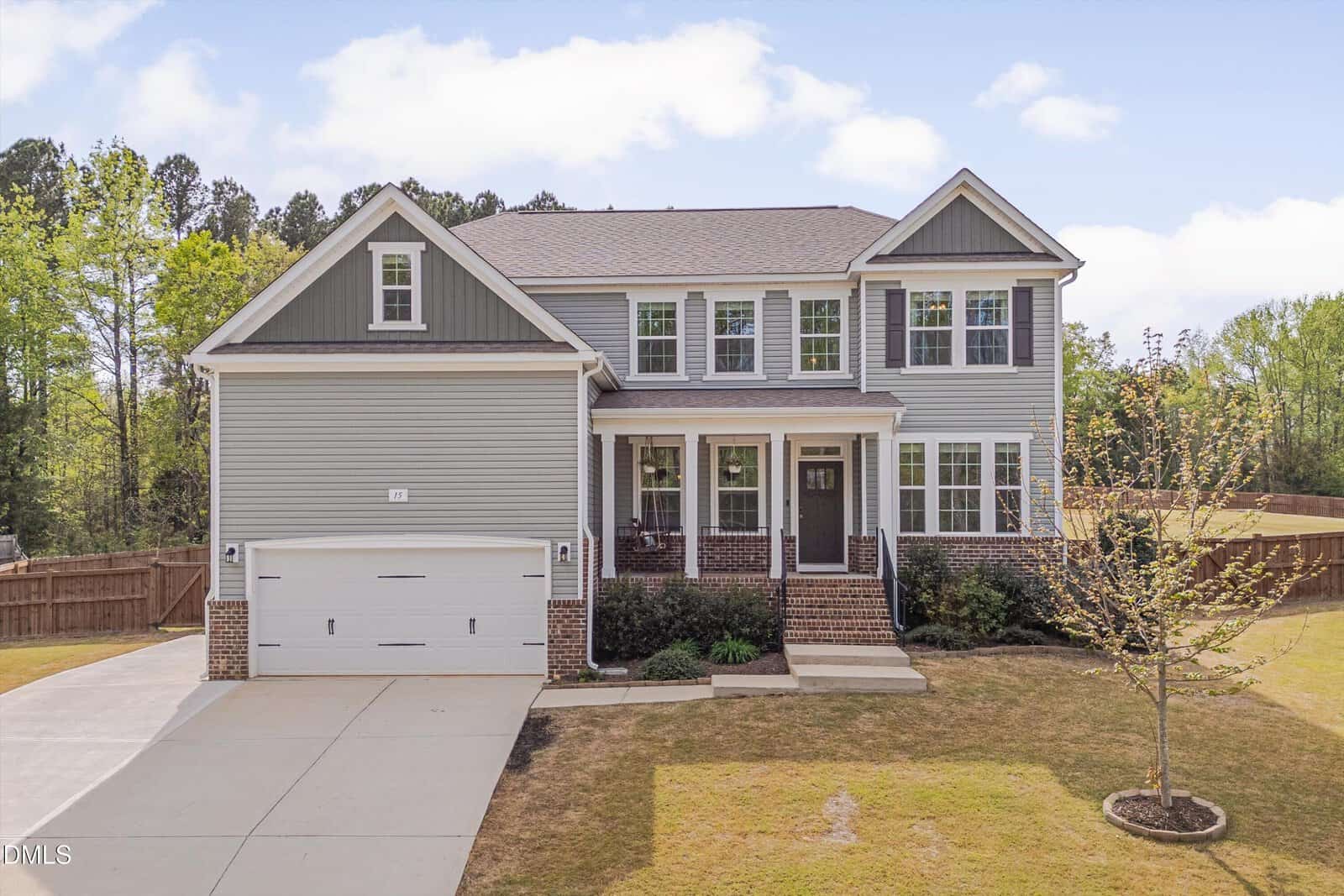 Two-story suburban house at 15 Oxer Drive in Youngsville, featuring light gray siding, white trim, and brick accents. Includes an attached two-car garage, front porch with columns, and a small tree in the front yard. Concrete driveway and fenced backyard provide added convenience and privacy.