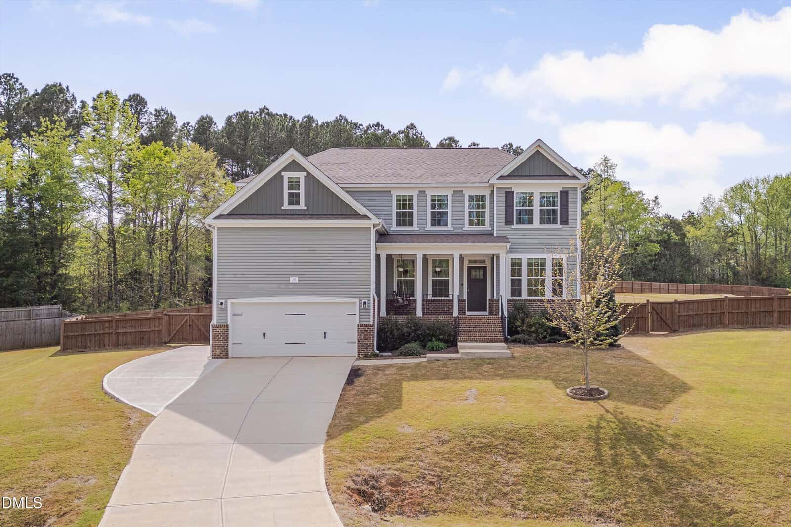 Two-story gray house with white trim, a front porch, attached two-car garage, and a curved driveway. The yard is spacious with a few small trees, surrounded by a wooden fence and bordered by woods.