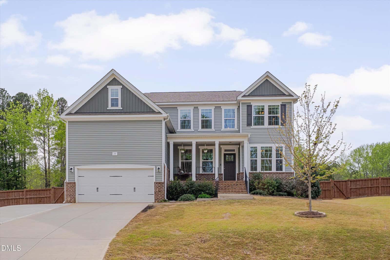 Two-story modern house with gray siding, white trim, a covered front porch, double garage, and a large driveway. The yard is grassy with a small tree, surrounded by a wooden fence and lots of greenery.