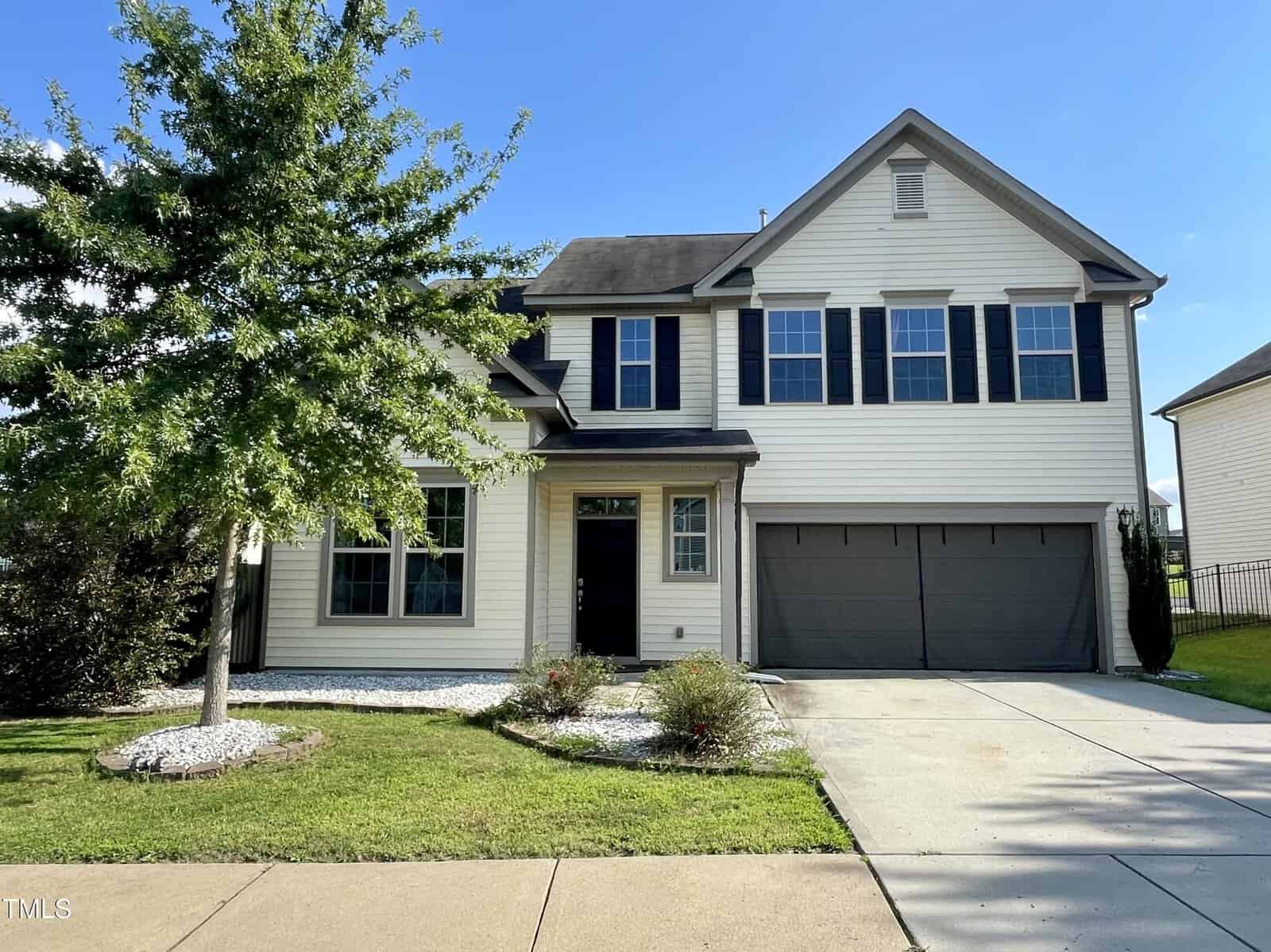 A two-story suburban house with cream siding, black shutters, and a double garage. A small tree and landscaped garden are in the front yard, bordered by a concrete driveway and walkway.