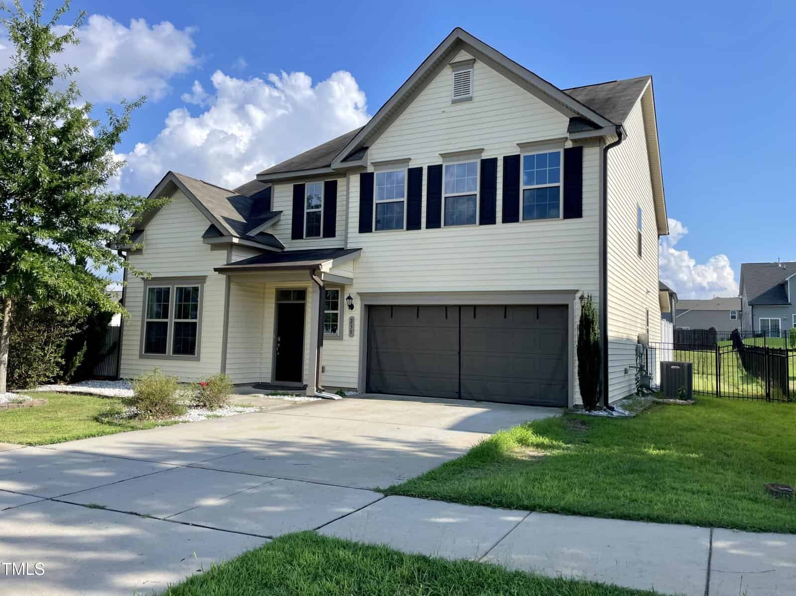 A two-story beige house with black shutters and a double garage sits on a concrete driveway, surrounded by green grass, a tree, and a partly cloudy sky in the background.