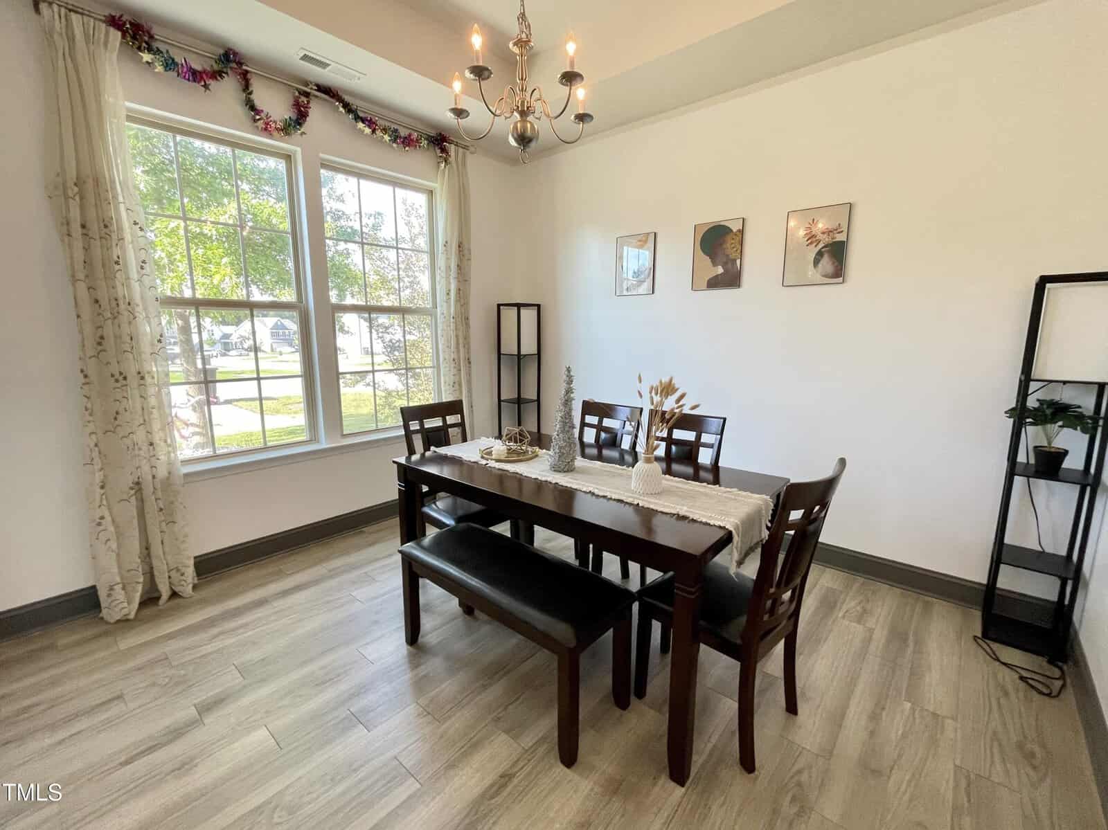 A dining room with a rectangular wooden table, four chairs, and a bench. The table has a runner and decorative items. There are two tall shelves, three framed pictures on the wall, a chandelier, and a large window with curtains.