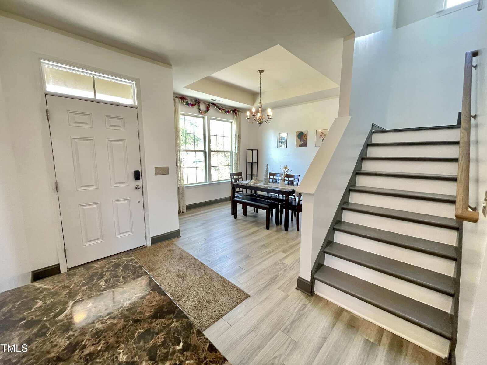 A view of a home's entryway with a white front door, stairs with a wooden handrail, and a dining area with a rectangular table, chairs, chandelier, and large window letting in natural light.