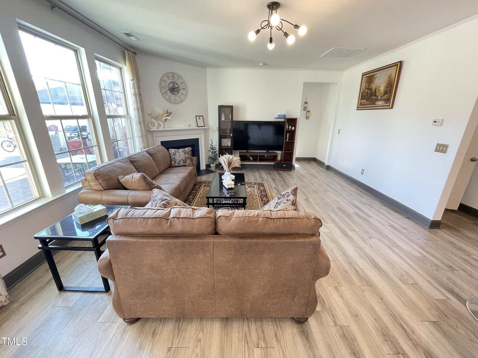 A spacious living room with light wood flooring, a brown couch and loveseat, a glass coffee table, a flat-screen TV on a stand, and large windows letting in natural light. A painting hangs on the white wall.